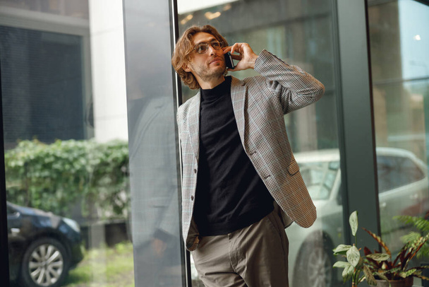Красивый бизнесмен разговаривает по мобильному телефону, стоит в коворкинге у окна и отворачивается. - Фото, изображение