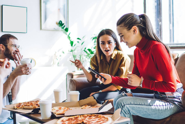 Cheerful woman in casual outfit browsing smartphone while smiling people eating delivered pizza and sitting on couch by table in stylish apartment - Foto, Imagen