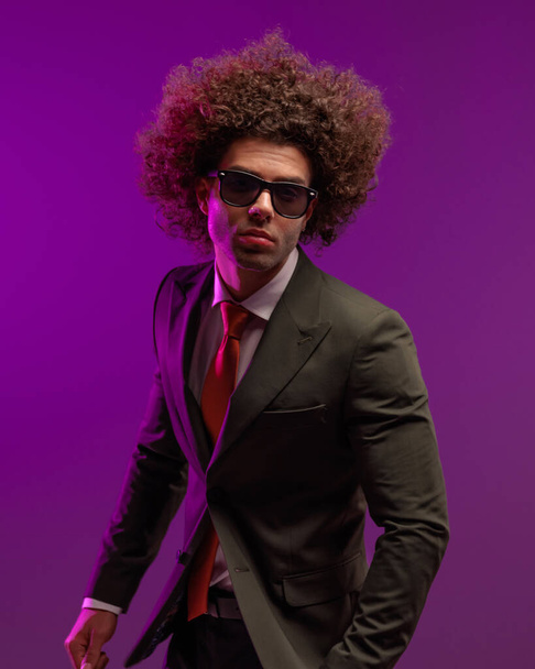 portret fajnego młodego biznesmena z długimi włosami w okularach przeciwsłonecznych i pozowanie przed fioletowym tle w studio - Zdjęcie, obraz
