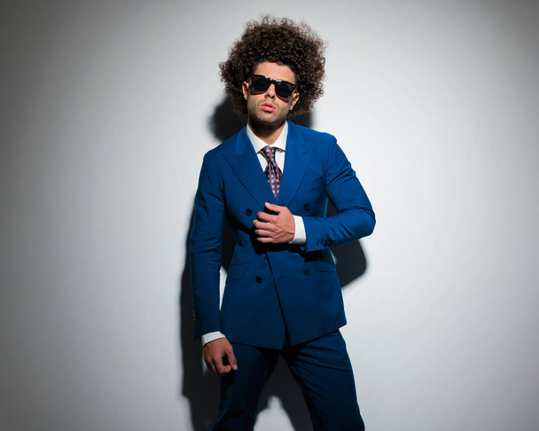 sexy high class man with afro hair arranging blue suit and posing in front of grey background  - Фото, изображение
