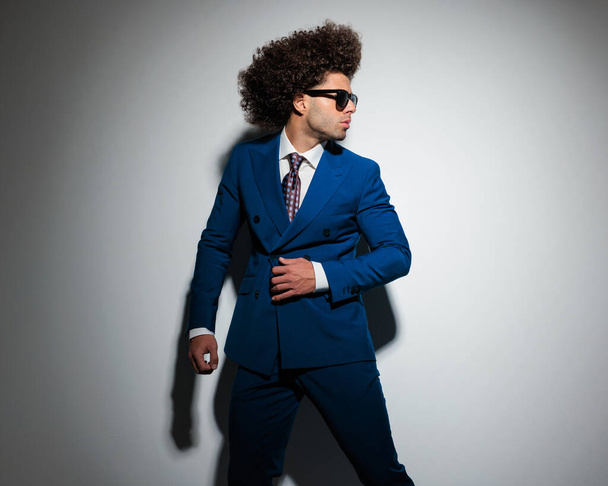 cool fashion man with afro hair and sunglasses looking to side and fixing blue suit in front of grey background  - Photo, Image