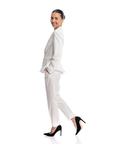 vue latérale d'une élégante jeune femme d'affaires en costume blanc heureuse, souriante et marchant les mains dans les poches devant un fond blanc - Photo, image