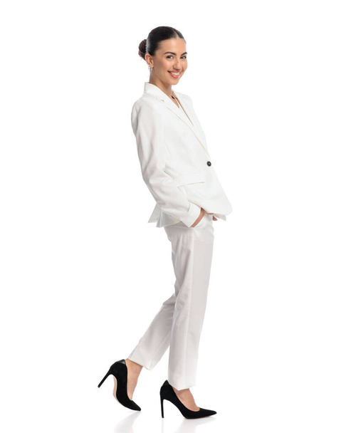 πλαϊνή άποψη του χαρούμενος νεαρό κορίτσι σε λευκό κοστούμι κρατώντας τα χέρια στις τσέπες, χαμογελώντας και περπατώντας μπροστά από το λευκό φόντο - Φωτογραφία, εικόνα
