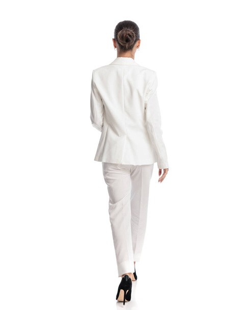 rear view of elegant fashion model with bun hair in elegant white suit walking in front of white background  - Photo, Image