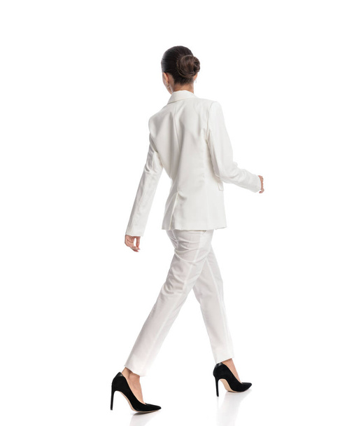 πλαϊνή άποψη της ελκυστικής γυναίκας με τα μαλλιά κουλούρι σε λευκό κοστούμι περπάτημα και ψάχνει για πλευρά μπροστά από το λευκό φόντο  - Φωτογραφία, εικόνα