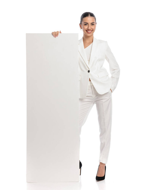 pewna siebie kobieta w białym garniturze pokazuje i prezentuje pustą deskę stojąc z ręką w kieszeni i uśmiechając się na białym tle  - Zdjęcie, obraz