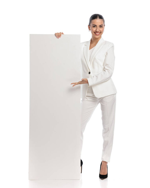 image complète du corps de la jeune femme élégante en costume blanc montrant tableau blanc vide et souriant devant fond blanc en studio - Photo, image