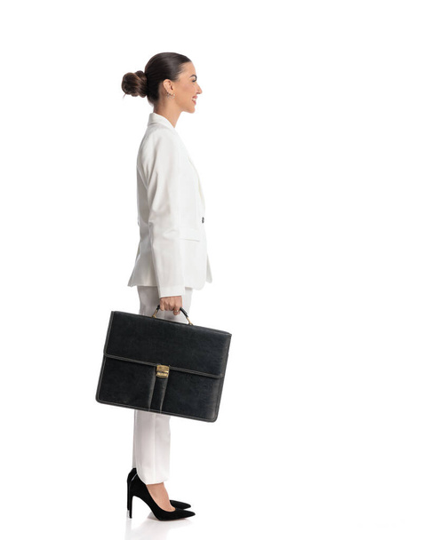 side view picture of sexy elegant girl in white suit with bag looking to side and smiling while standing in line in front of white background  - Photo, Image
