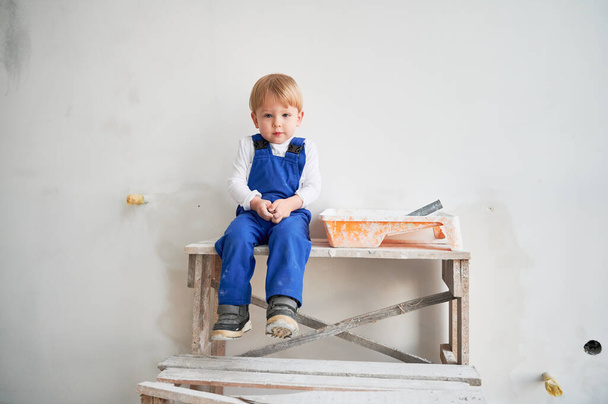 Cute kid construction worker sitting on wooden table against white wall in apartment under renovation. Cheerful little boy wearing safety helmet and work overalls while playing at home. - Foto, Imagem