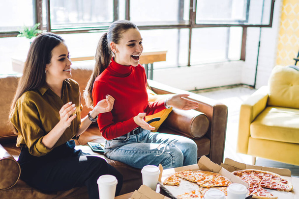 Веселые люди смеются и жестикулируют, сидя на диване за столом с чашками на вынос и доставляя пиццу домой при дневном свете - Фото, изображение