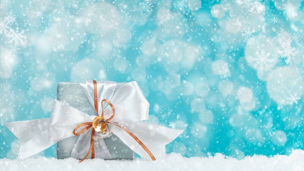 白と金のリボンボウでブルーシルバーペーパーに包まれたギフトボックス. 青い背景,雪とボケのライト. クリスマスと新年の贈り物,ボクシングデー. - 写真・画像