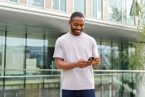 Африканский американец с сенсорным экраном смартфона печатает страницу прокрутки на городской улице в городе. Парень с сотовым телефоном, который пользуется интернетом через социальные сети. Зависимость от мобильных телефонов - Фото, изображение