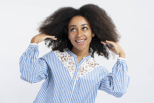 Ritratto di bella donna afroamericana positiva che fissa i capelli ricci distogliendo lo sguardo isolato su sfondo bianco. Concetto di cura dei capelli, procedure cosmetiche, trattamento - Foto, immagini