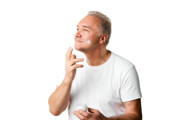 Porträt eines Mannes mittleren Alters, 45 Jahre alt, der vor weißem Studiohintergrund Hautpflegecreme auf das Gesicht aufträgt. Konzept der natürlichen Schönheit, Hautpflege, Gesundheitsfürsorge, Anti-Aging-Behandlung. Kopierraum. Anzeige - Foto, Bild