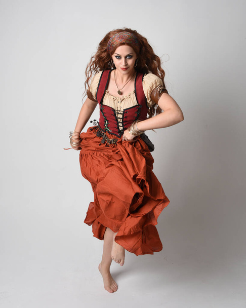 Ortaçağ bakiresi, falcı kostümü giymiş güzel kızıl saçlı bir kadının tam boy portresi. Dans hareketleriyle ayakta durmak, etek sallamak. Stüdyo arka planında izole. - Fotoğraf, Görsel