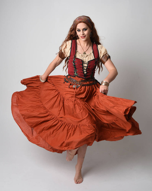 Ortaçağ bakiresi, falcı kostümü giymiş güzel kızıl saçlı bir kadının tam boy portresi. Dans hareketleriyle ayakta durmak, etek sallamak. Stüdyo arka planında izole. - Fotoğraf, Görsel