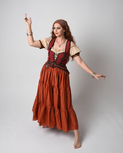 Полный портрет красивой рыжеволосой женщины в средневековой девичьей одежде, костюм гадалки. Стоя в позе с танцевальными жестами, закручивая юбку. изолированные на студийном фоне. - Фото, изображение
