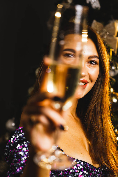Portret czarującej białej kobiety w błyszczącej niebieskiej sukience w pobliżu choinki z kieliszkiem szampana.Koncepcja noworoczna i świąteczna - Zdjęcie, obraz