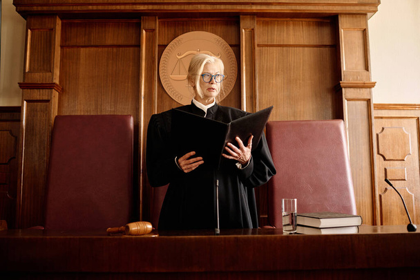 Впевнена зріла блондинка-жінка-суддя з відкритою текою, що стоїть за столом у залі суду і оголошує вирок підозрюваному після судового засідання - Фото, зображення