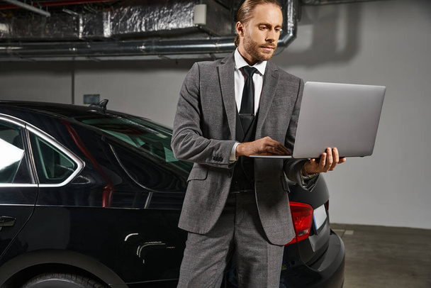 przystojny mężczyzna w inteligentnym garniturze pracuje na laptopie na parkingu w pobliżu jego samochodu, koncepcja biznesu - Zdjęcie, obraz