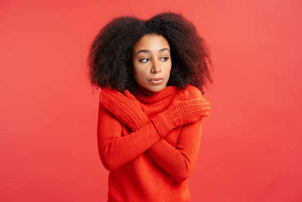Porträt einer aufgebrachten schönen Afroamerikanerin mit lockigem Haar, die rote Handschuhe trägt und isoliert auf rotem Hintergrund wegschaut. Winterkonzept - Foto, Bild