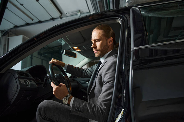 όμορφος γενειοφόρος άνδρας με κόκκινα μαλλιά σε έξυπνο κομψό κοστούμι πίσω από το τιμόνι του αυτοκινήτου του, επιχείρηση - Φωτογραφία, εικόνα