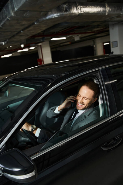 cheerful appealing man with beard and ponytail talking by phone behind steering wheel, business - Photo, Image
