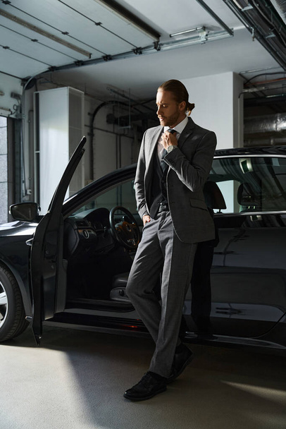 homme barbu attrayant en costume élégant posant à côté de sa voiture avec la main sur sa cravate, concept d'affaires - Photo, image
