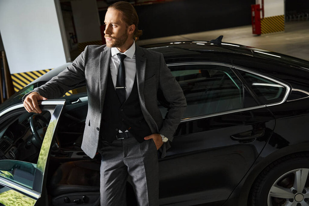 atraktivní elegantní profesionál v elegantním elegantním elegantním obleku pózuje v blízkosti svého auta s rukou v kapse, obchod - Fotografie, Obrázek