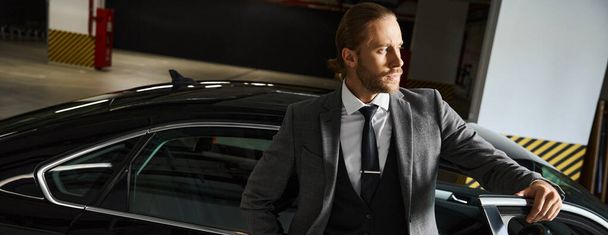 ελκυστικό γενειοφόρος άνδρας με έξυπνη στολή ποζάροντας κοντά στο αυτοκίνητό του και κοιτάζοντας μακριά, επιχειρηματική ιδέα, πανό - Φωτογραφία, εικόνα