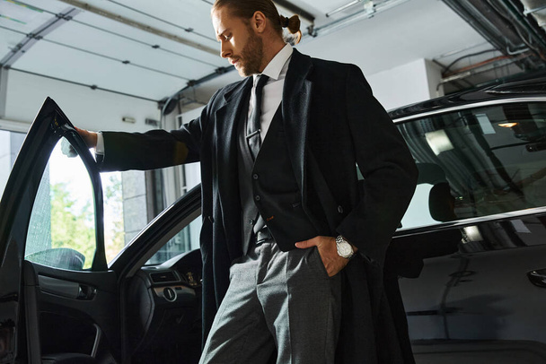 привлекательный элегантный мужчина в элегантной одежде позирует рядом со своим автомобилем с рукой в кармане, бизнес-концепция - Фото, изображение