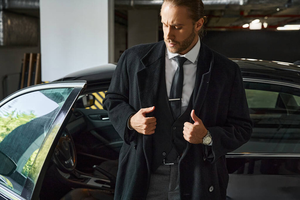 красивий рудий чоловік з хвостом у чорному розумному пальто, що позує на парковці, бізнес-концепція - Фото, зображення
