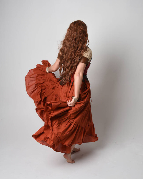 Ολο το μήκος πορτρέτο της όμορφης κοκκινομάλλας γυναίκας που φοράει μια μεσαιωνική παρθένα, κοστούμι μάντη. Στέκεται πίσω θέα, περπατώντας μακριά. απομονωμένο σε φόντο στούντιο. - Φωτογραφία, εικόνα