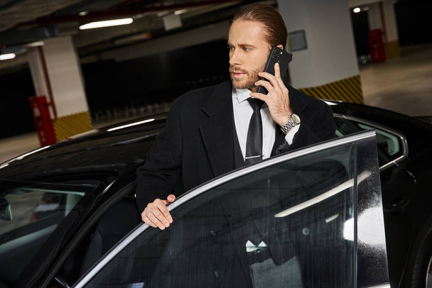 appealing bearded man in smart black attire talking by phone next to his car, business concept - Photo, Image
