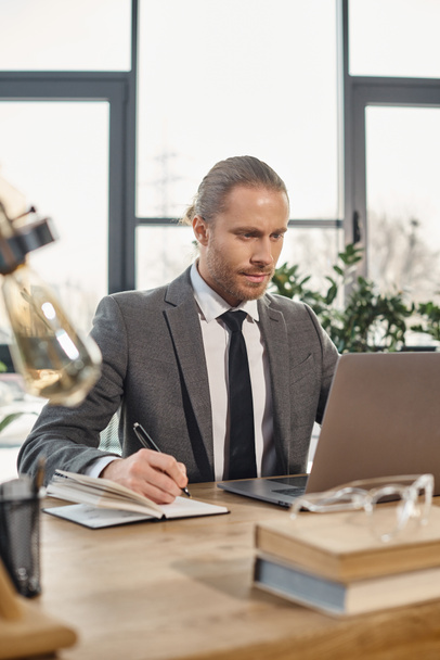 konzentrierter Geschäftsmann, der im Notizbuch schreibt und am Arbeitsplatz im modernen Büro auf den Laptop blickt - Foto, Bild