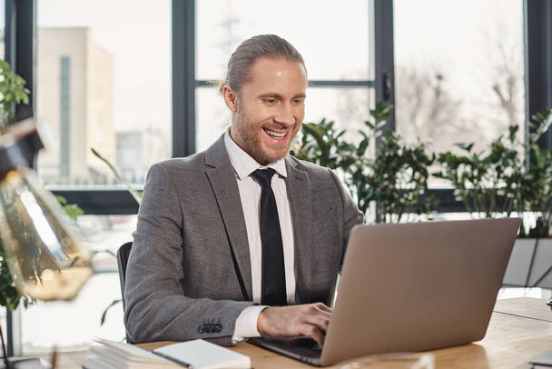 iloinen ja tyylikäs liikemies harmaassa puvussa kirjoittamalla kannettavan tietokoneen työskennellessään modernissa toimistossa - Valokuva, kuva