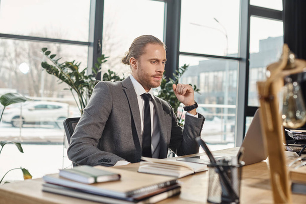 pensativo hombre de negocios en ropa formal mirando a la computadora portátil mientras está sentado en el lugar de trabajo en la oficina moderna - Foto, imagen