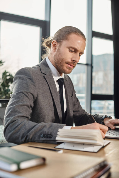 keskittynyt liikemies harmaassa puvussa kirjoittaa kannettavaan lähelle kannettavaa nykyaikaisessa työympäristössä - Valokuva, kuva
