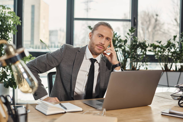 серьезный стильный бизнесмен сидит на рабочем месте рядом с ноутбуком и смотрит в камеру в офисе - Фото, изображение