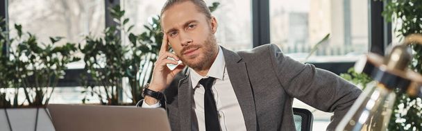 задумчивый бизнесмен в сером костюме сидит на рабочем месте рядом с ноутбуком и смотрит в камеру, баннер - Фото, изображение