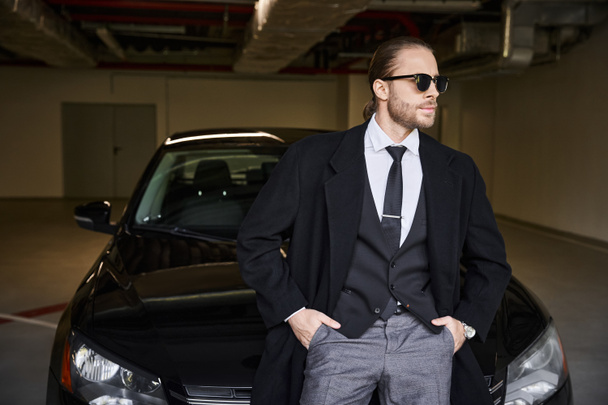 attrayant homme barbu avec des lunettes de soleil posant près de sa voiture avec les mains dans les poches, concept d'affaires - Photo, image