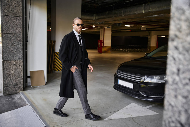 όμορφος κομψός επιχειρηματίας με μαύρο παλτό με γυαλιά ηλίου περπατώντας και κοιτάζοντας την κάμερα - Φωτογραφία, εικόνα