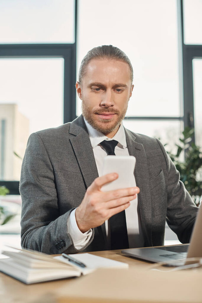 doordachte zakenman in grijs pak zit naast laptop op het bureau en kijkt naar smartphone - Foto, afbeelding