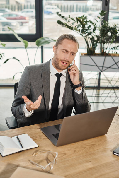 homem de negócios sério olhando para laptop durante a conversa no smartphone no local de trabalho no escritório - Foto, Imagem