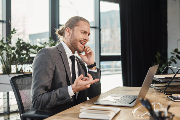 hermostunut liikemies osoittaa kannettavan tietokoneen puhuessaan matkapuhelimella työpaikalla toimistossa - Valokuva, kuva