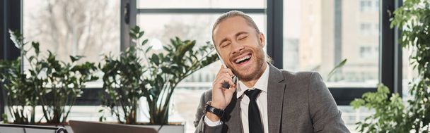 przepełniony sukcesem biznesmen śmiejący się podczas rozmowy na smartfonie w biurze, baner - Zdjęcie, obraz