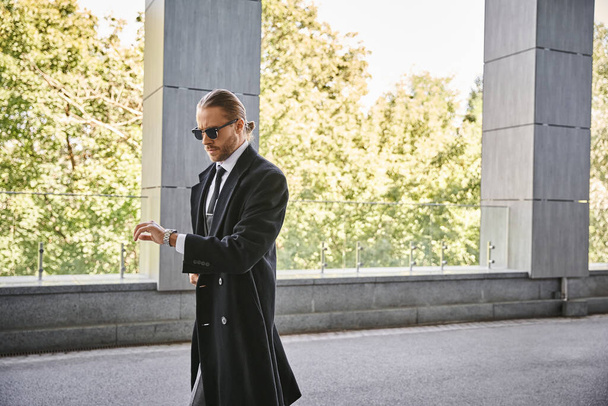 pohledný podnikatel s nádherným elegantním stylem se slunečními brýlemi při pohledu na hodinky - Fotografie, Obrázek