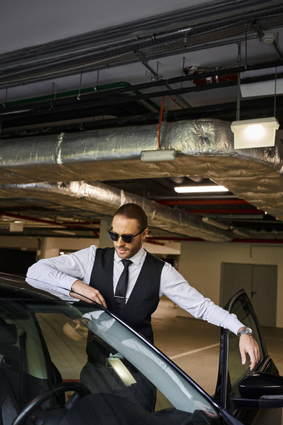 atractivo hombre barbudo con pelo rojo en chaleco negro con gafas de sol posando cerca de su coche, negocio - Foto, imagen