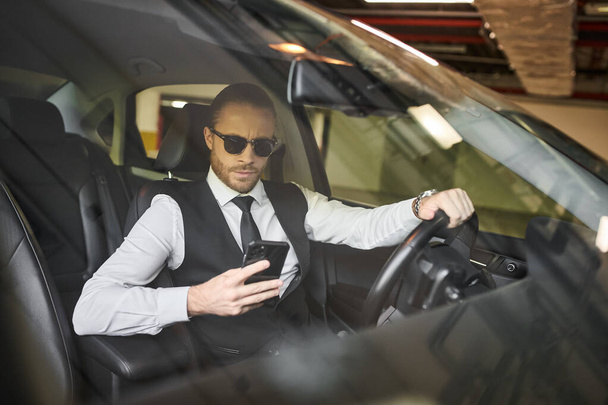 przystojny uroczy biznesmen w czarnej kamizelce w okularach przeciwsłonecznych patrząc na telefon za kierownicą - Zdjęcie, obraz