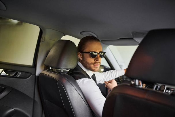 όμορφος γενειοφόρος επιχειρηματίας με γυαλιά ηλίου κοιτάζοντας κάμερα, ενώ πίσω από το τιμόνι - Φωτογραφία, εικόνα
